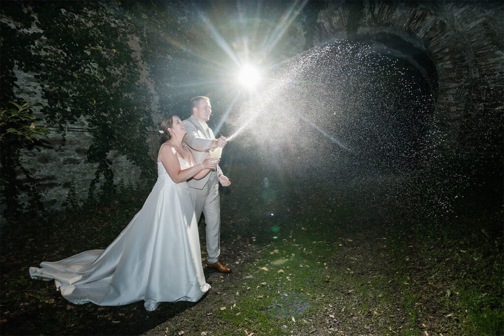 Braut und Bräutigam spritzen lachend eine Champagnerflasche bei ihrer Hochzeit, während das Licht die funkelnden Tropfen im nächtlichen Grugapark beleuchtet