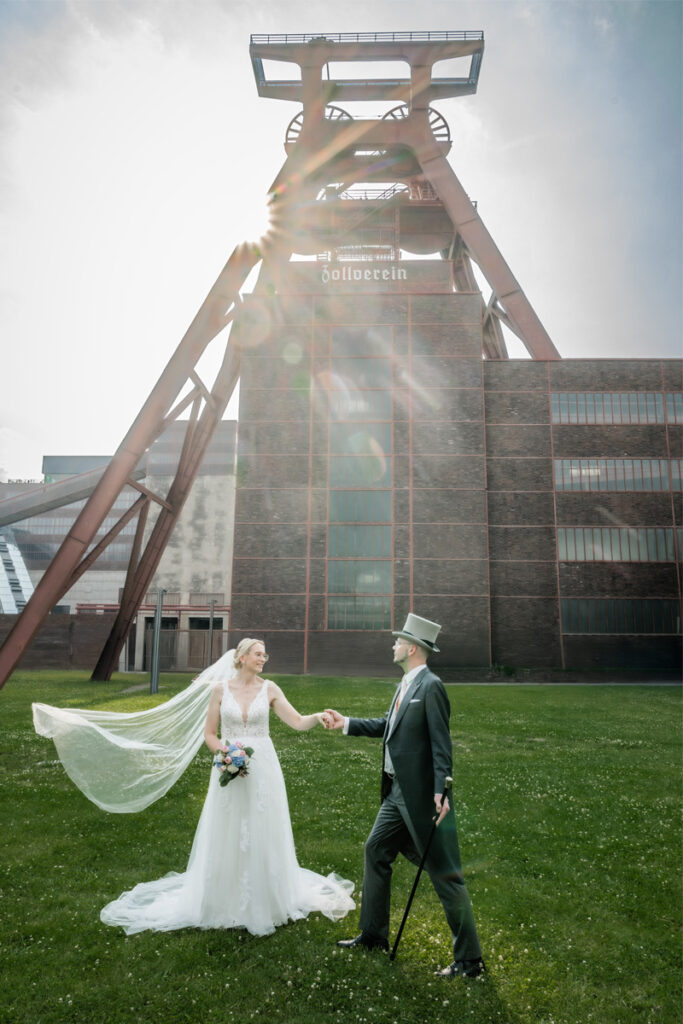Braut und Bräutigam vor der Zeche Zollverein in Essen – romantisches Hochzeitspaar mit wehendem Schleier und Sonnenlicht im Hintergrund