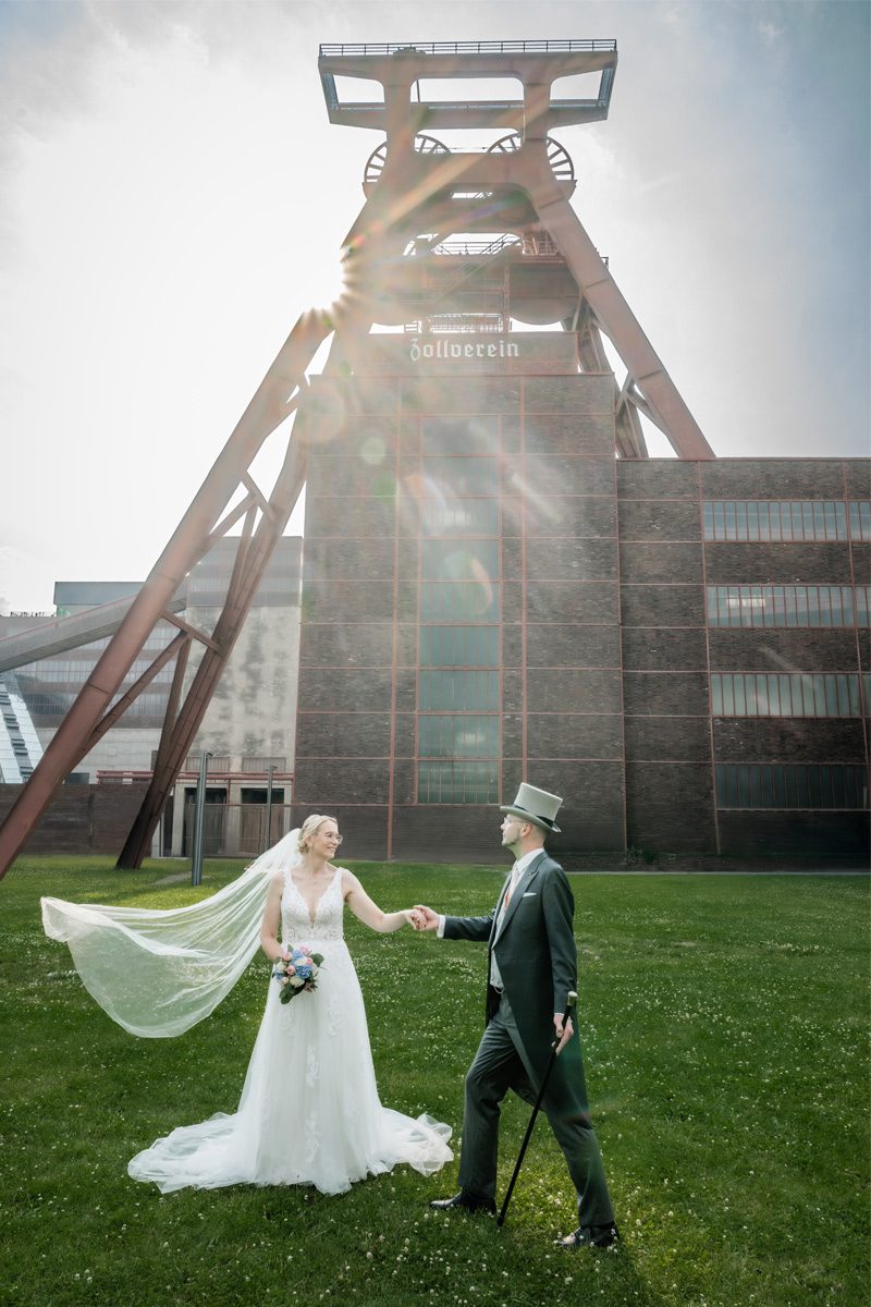 Braut und Bräutigam vor der Zeche Zollverein in Essen – romantisches Hochzeitspaar mit wehendem Schleier und Sonnenlicht im Hintergrund