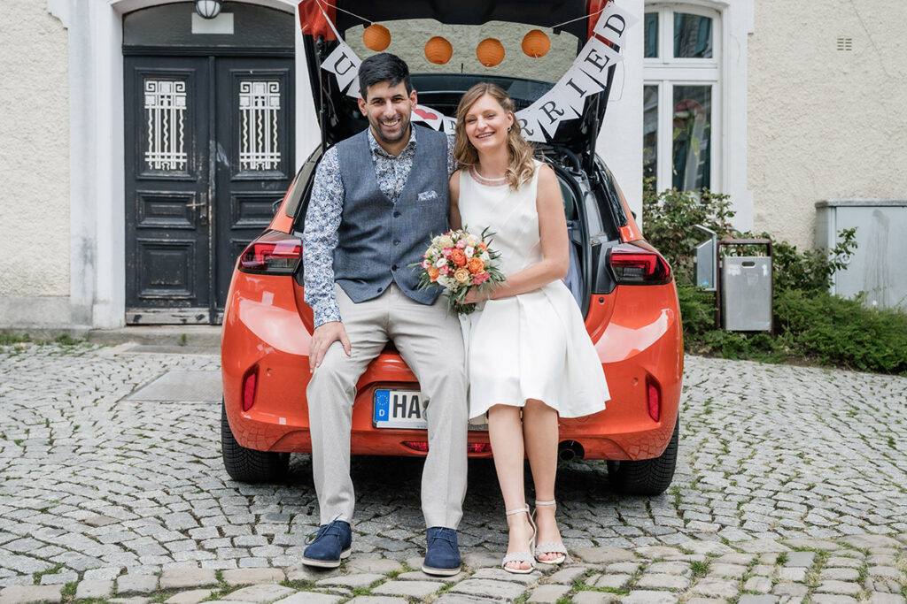 Modernes Brautpaar sitzt im Kofferraum eines orangefarbenen Autos mit 'Just Married'-Banner vor dem Romantikhotel Neuhaus, Hochzeitsfotografie Andreas Zabel