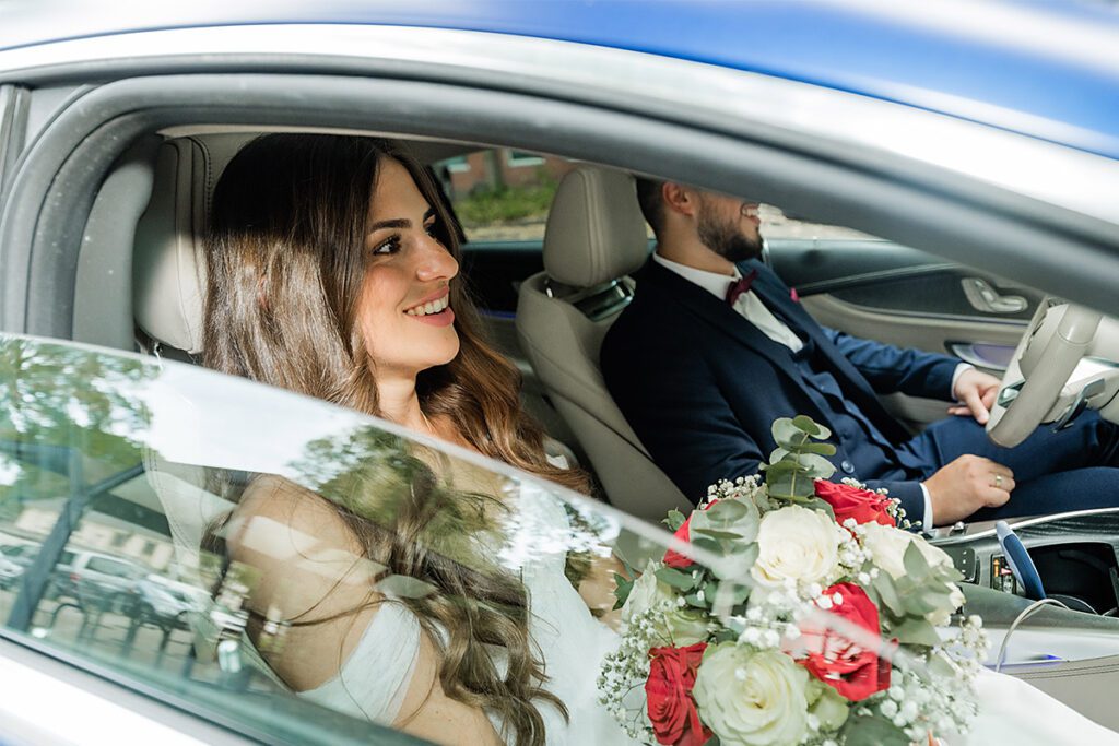 Fahrt zur Hochzeitslocation in Essen: Glückliche Braut mit Rosenstrauß auf dem Beifahrersitz im blauen Auto