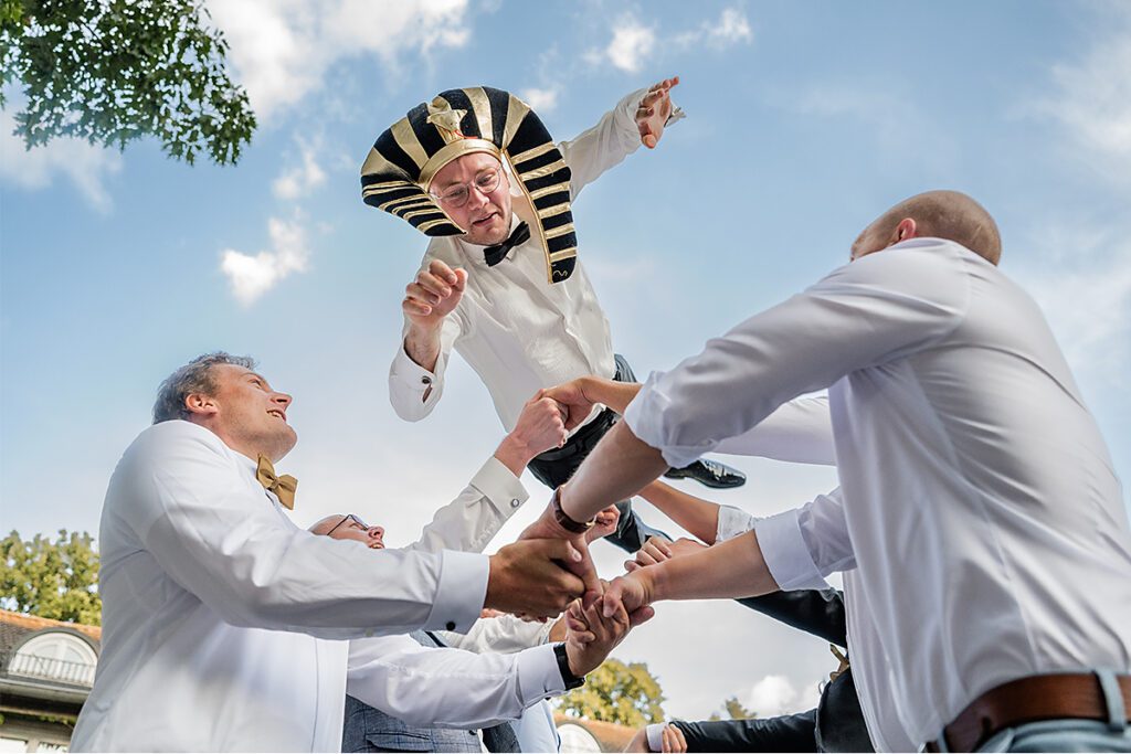 Lustiges Hochzeitsspiel in Essen: Bräutigam mit Pharao-Mütze wird von seinen Gästen jubelnd in die Luft geworfen