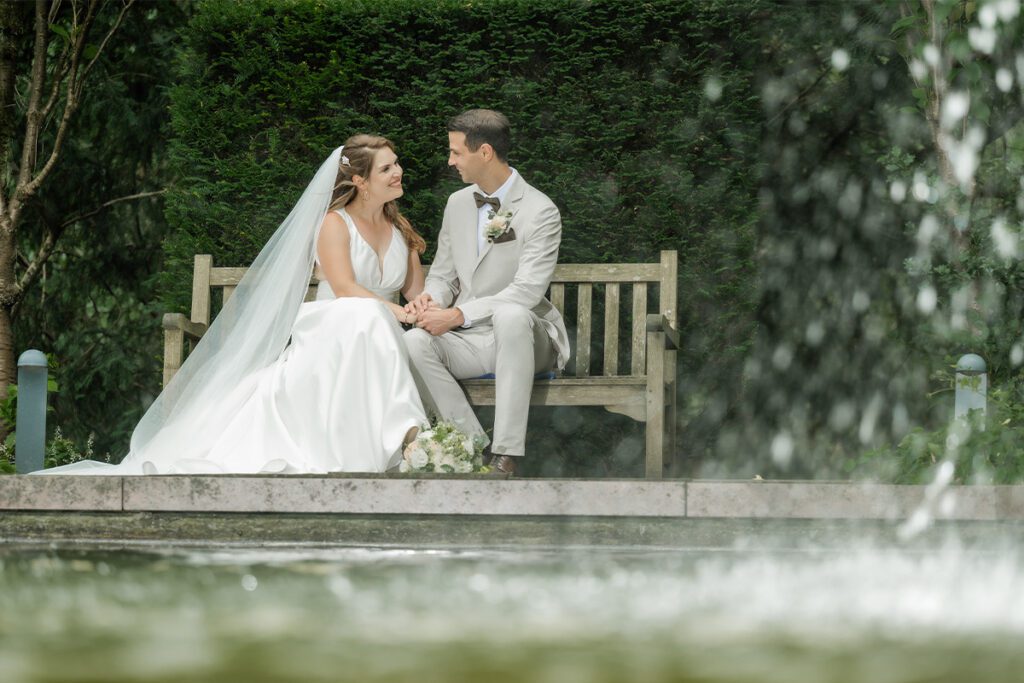 Braut im weißen Hochzeitskleid mit Schleier und Bräutigam im beigen Anzug sitzen Hand in Hand auf einer Parkbank, fotografiert durch unscharfe Wasserspritzer im Vordergrund