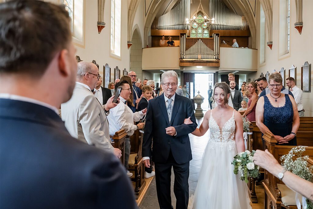 Blick über die Schulter des wartenden Bräutigams auf die eingreifende Braut im weißen A-Linien-Kleid mit Brautstrauß, die am Arm ihres Vaters durch den Mittelgang der Kirche geht